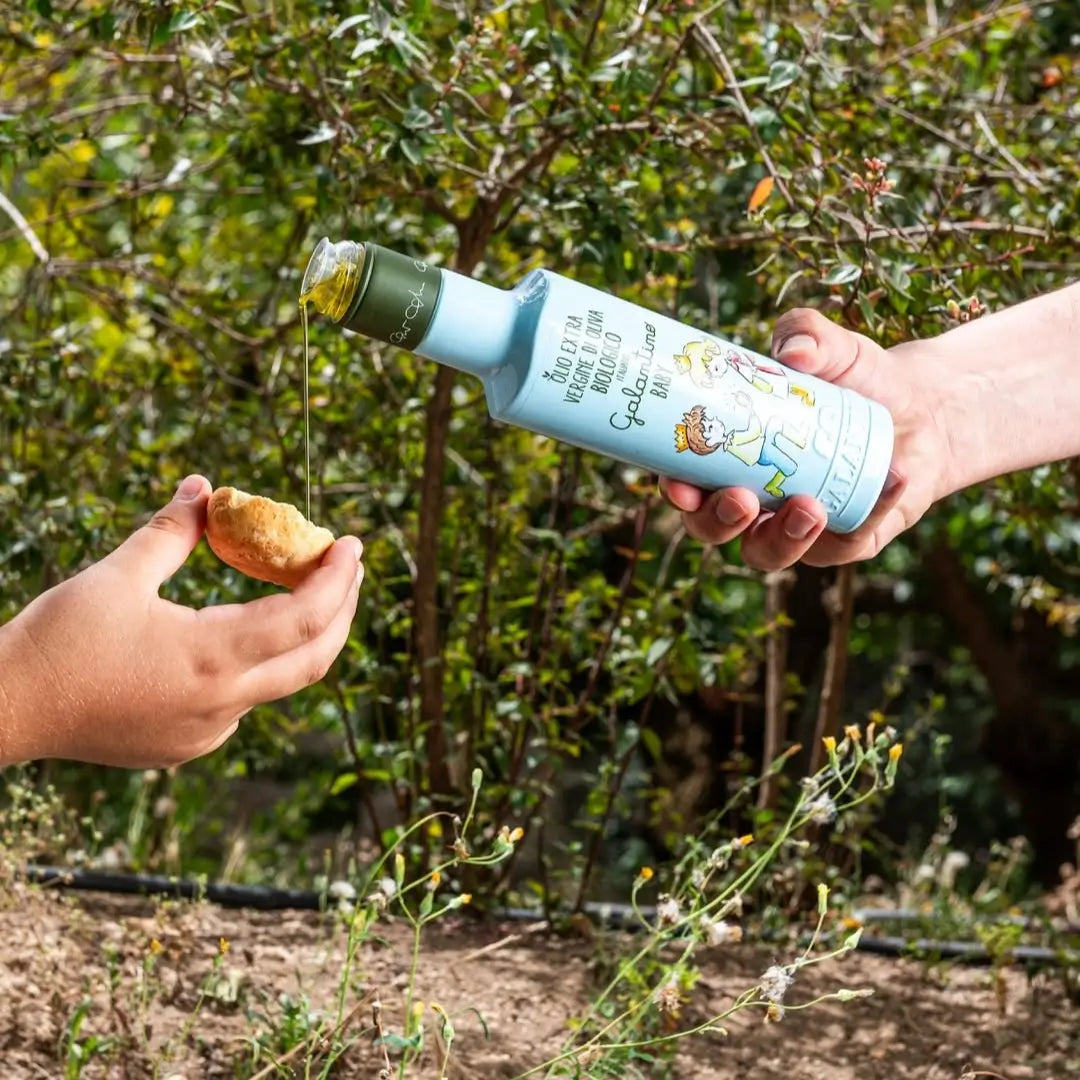 Olijfolie wordt uit een lichtblauwe fles op brood gegoten in een groene buitenomgeving, met de tekst 'Good things take time' op het etiket